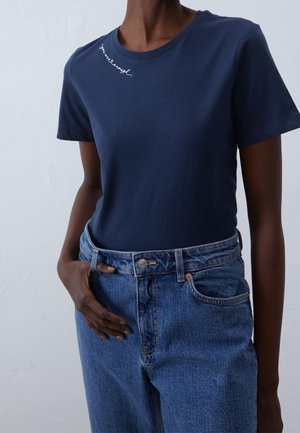 Osoba v tmavě modrém tričku s bílým kurzívním nápisem poblíž límce a v modrých džínách s vysokým pasem, ruka opřená o bok.