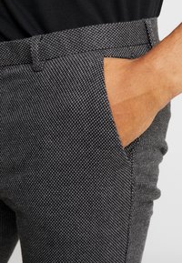 Šedé vzorované kalhoty s texturovanou látkou, vybavené postranními kapsami a plochým předním pasem. Detailní pohled ukazuje detailní tkaní.