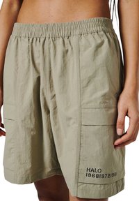 Licht khaki cargoshorts met elastische tailleband en bedrukte tekst "HALO 196819721981" op het onderste rechterzakje, gedragen door een persoon.