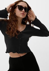 Schwarzes geripptes Crop-Top mit V-Ausschnitt und gewelltem Saum, mit einer Knopfleiste an der Vorderseite. Kombiniert mit strukturierten schwarzen High-Waist-Hosen.