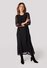 Zwarte jurk met een ronde halslijn, doorschijnende lange mouwen en een gestrikt taille-detail. Glad textuur met een gelaagd ontwerp dat tot de zoom reikt.