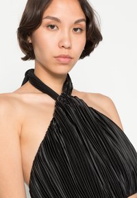 Top halter preto plissado com decote torcido, feito de um tecido suave. O padrão apresenta plissados verticais e uma silhueta ajustada.