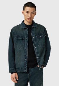 Tummansinisestä gradienttidesignista valmistettu farkkutakki, jossa on kaksi rintataskua, napit ja klassinen kaulus. Päällä musta villapaita.