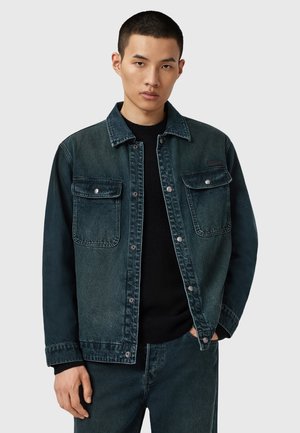 Chaqueta de mezclilla en un diseño de degradado azul oscuro con dos bolsillos en el pecho, botones de presión y un cuello clásico. Lleva puesta una sudadera negra.