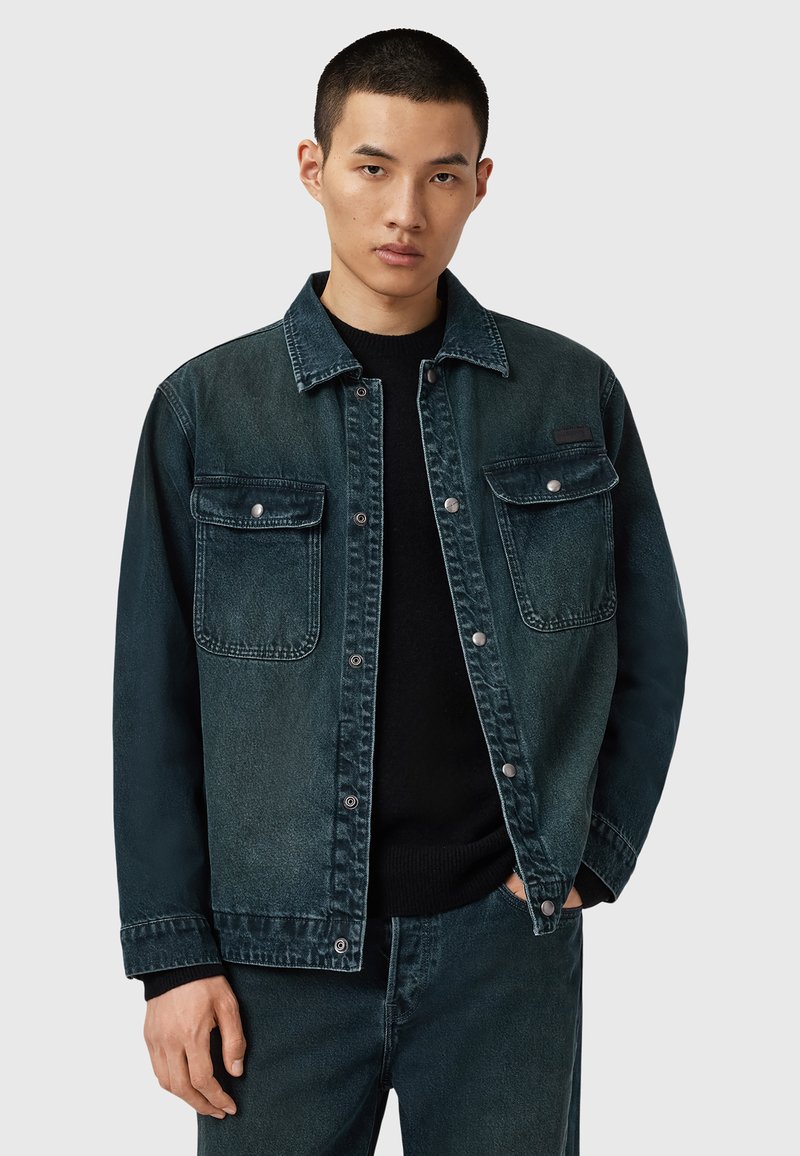 Tummansinisestä gradienttidesignista valmistettu farkkutakki, jossa on kaksi rintataskua, napit ja klassinen kaulus. Päällä musta villapaita.