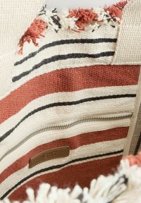 Intérieur d'un sac fourre-tout tissé avec des rayures rouge, noir et crème, poche zippée et accents texturés. Tissu doux avec des détails à franges.