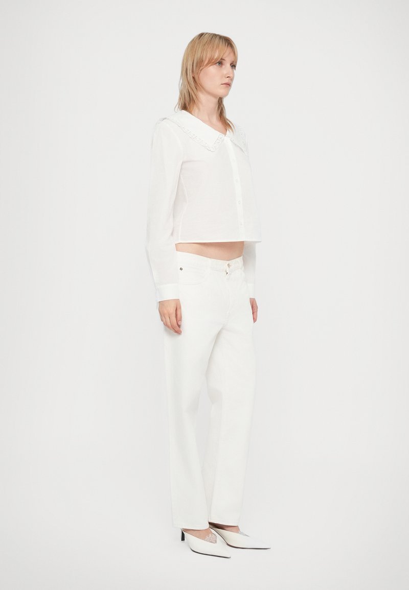 Weißes, kurz geschnittenes Hemd mit einer Spitzenkragen, kombiniert mit weißen, hochgeschnittenen Jeans und weißen, spitzen High Heels.