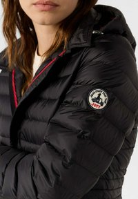 Chaqueta acolchada negra con patrón de acolchado, acentos rojos a lo largo de la cremallera y el cuello, con un parche redondo de logo en la manga izquierda.