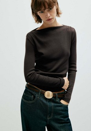 Jeune personne aux cheveux courts et bruns portant un haut noir ajusté à manches longues et un jean bleu avec une ceinture marron et une boucle ovale.