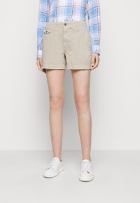 Žena v béžových šortkách, modro-růžové kostkované košili na knoflíky a bílých teniskách stojící před jednobarevným šedým pozadím.