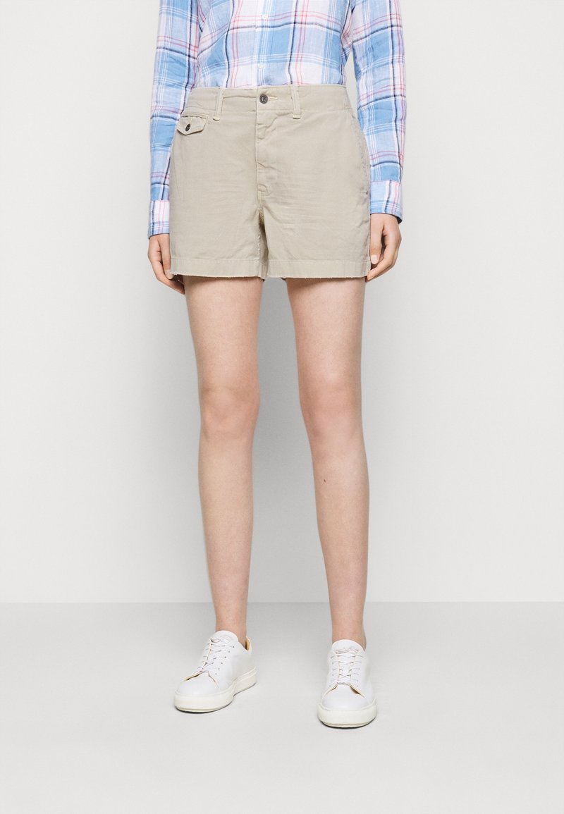 Žena v béžových šortkách, modro-růžové kostkované košili na knoflíky a bílých teniskách stojící před jednobarevným šedým pozadím.