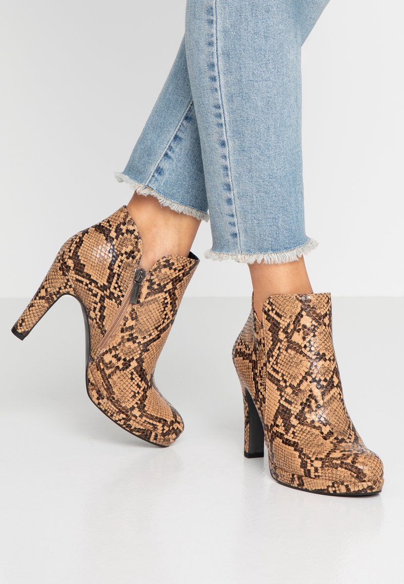 Bottes à talons hauts à motif peau de serpent montant jusqu'à la cheville, portées avec un jean bleu clair à ourlet effiloché, montrant un détail de fermeture éclair latérale.