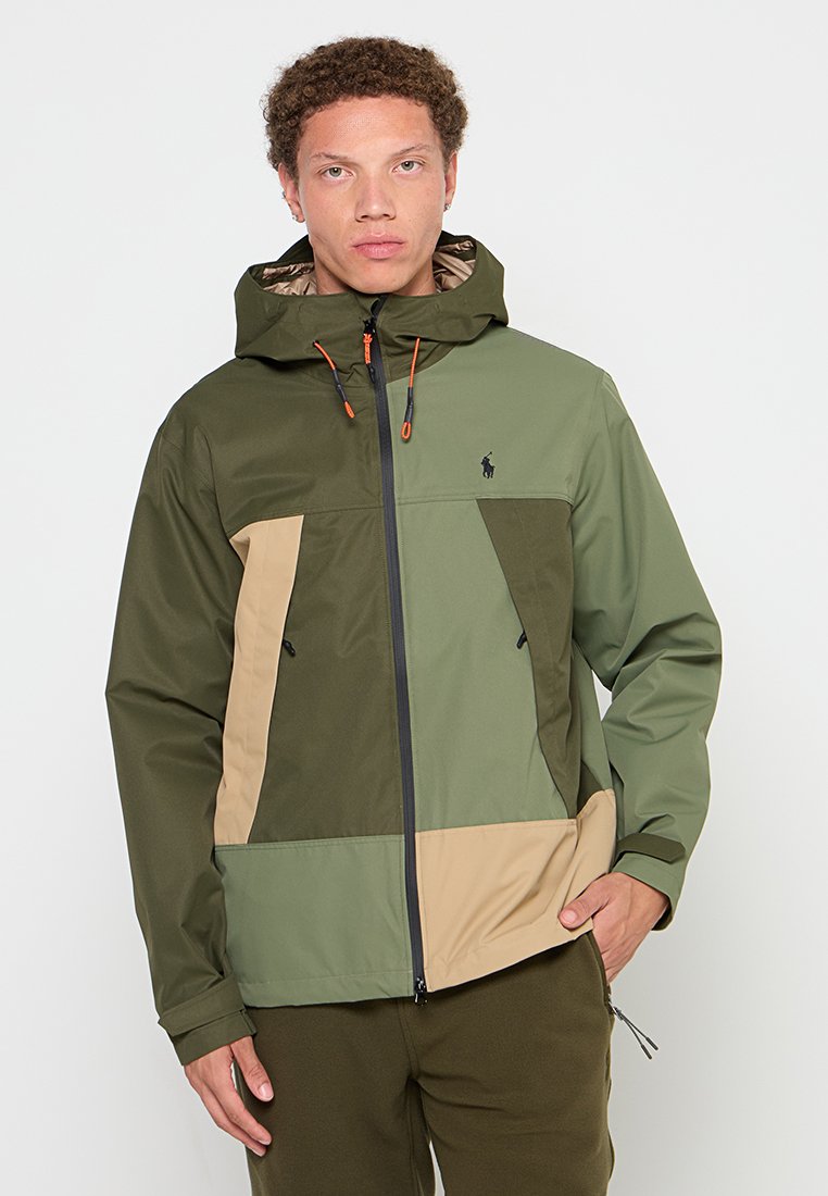 Giacca color-block in verde oliva e beige, realizzata in un tessuto leggero. Presenta un cappuccio, una zip frontale e cordini regolabili.