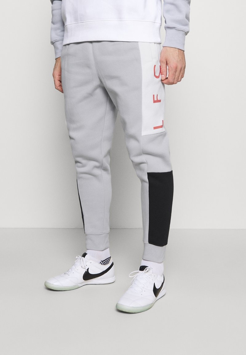 Jogging gris clair avec panneaux latéraux blancs et lettres rouges, patchs noirs sur le bas des jambes, porté avec des baskets Nike blanches et des chaussettes blanches.