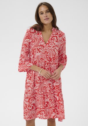 Kobieta ubrana w czerwioną sukienkę do kolan z białymi wzorami paisley, z rękawami o długości trzy czwarte i dekoltem w serek, stojąca na jednolitym tle.
