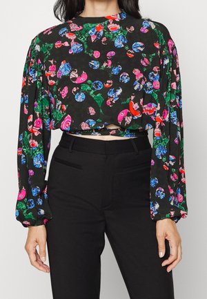 Žena nosí dlouhý rukáv krátký top s barevnými květinovými a abstraktními vzory, doplněný o černé kalhoty s vysokým pasem.