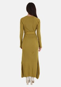 Abito lungo a maglia verde oliva con righe, vita con cintura, maniche lunghe e spacchi laterali, indossato con stivali bianchi con tacco quadrato.
