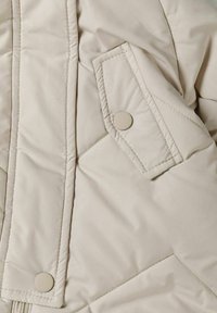 Detaliu al unei jachete puffer bej, având textură matlasată, butoane cu clapă și un buzunar lateral. Material sintetic neted cu un luciu subtil.