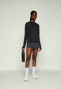 Žena v černém dlouhém rukávu, vzorovaných šortkách, bílých Nike ponožkách a botách, držící černou Nike láhev na vodu, stojící na jednobarevném pozadí.