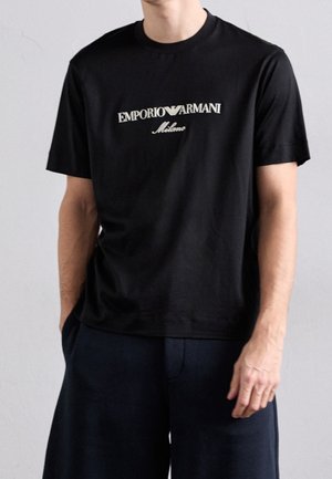 Mężczyzna ubrany w czarną koszulkę z krótkim rękawem Emporio Armani Milano i ciemne szorty, stoi z jedną ręką w kieszeni na szarym tle.