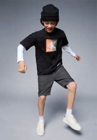 Schwarzes Kurzarm-T-Shirt mit grafischem Design, kombiniert mit grauen Shorts und weißen Sneakern. Das Model trägt eine schwarze Mütze und ein weißes Unterhemd.
