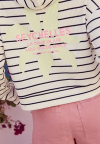 Wit met marineblauwe gestreepte capuchontrui met een groene blaadjesafbeelding en roze tekst. Gecombineerd met lichtroze korte broek. De tekst bevat plaatsnamen.