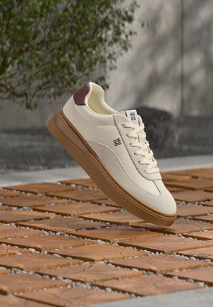 Sneaker color crema con suola marrone e dettaglio sul tallone, marchiata "Hugo", sospesa sopra un pavimento esterno in legno e ciottoli a motivo.