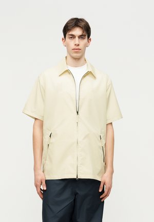 Jauns vīrietis ar īsiem tumšiem matiem valkā krēma krāsas īsroku kreklu ar rāvējslēdzēju virs baltas krekla un tumšām biksēm, stāvot pret vienkrāsainu fonu.