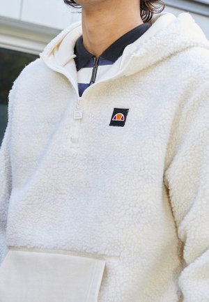 Haut à capuche en polaire blanche avec une poche kangourou et un patch logo noir. Matériau texturé et doux avec une coupe décontractée et un col superposé.