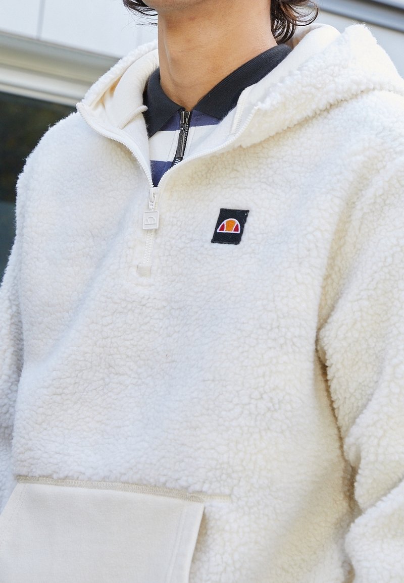 Vitt fleece hoodie med kvartdragkedja, känguruficka och svart logotyp-patch. Texturerat, mjukt material med avslappnad passform och lager-kraja.