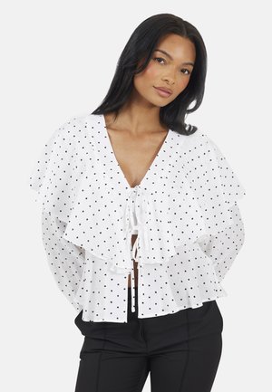 Kobieta o długich czarnych włosach ubrana w białą bluzkę w kropki z falbanianym kołnierzem i wiązaniami z przodu, w połączeniu z czarnymi spodniami, na jednolitym tle.