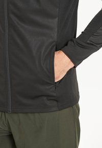Zwarte sporttop met rits, gemaakt van textuurstof, met zijzakken en versterkte naden, in combinatie met groene broeken, wat een sportieve uitstraling benadrukt.