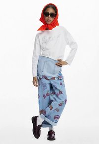 Sudadera blanca recortada con bordado "Delight", pantalones vaqueros azules con patrones florales, bufanda roja y gafas de sol negras. Zapatos marrones.