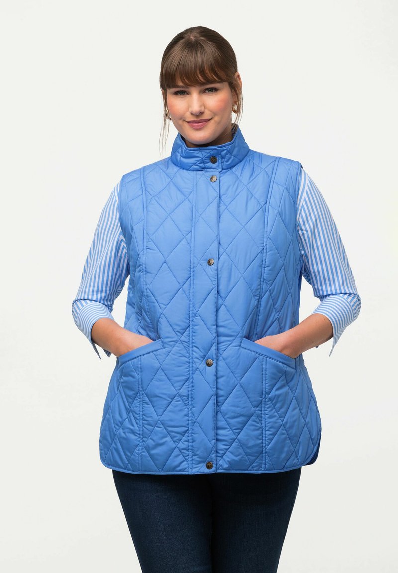 Blå vadderad väst med hög krage, tryckknappar och framfickor; bärs över en randig skjorta. Tillverkad av lätt, vadderat material.