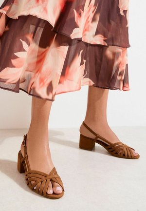 Woman wearing brown suede open-toe block heel sandals and a flowy floral skirt with pink and brown tones.
