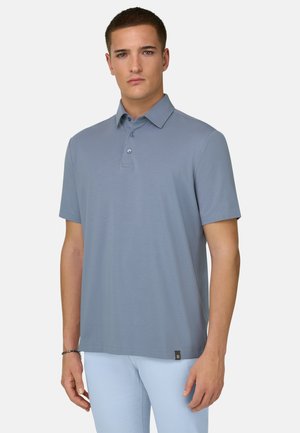 Jeune homme portant un polo bleu clair à manches courtes avec des boutons et un pantalon de couleur claire, debout devant un fond uni.