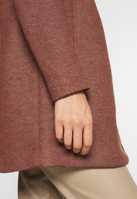 Tela marrón texturizada de un abrigo largo, con una manga acampanada y una mano posicionada en la costura, combinada con unos pantalones beige claros.