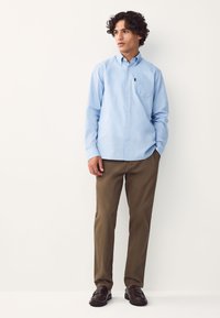 Camicia azzurro chiaro con bottoni e taschino, abbinata a pantaloni marroni e mocassini scuri, con taglio dritto e tessuto liscio.