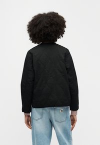 Giacca trapuntata nera con scollo rotondo e maniche lunghe, abbinata a pantaloni di jeans azzurri. Presenta dettagli di cuciture sottili.