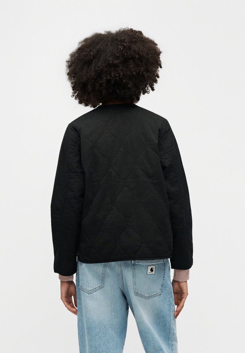 Zwarte doorgestikte jas met een ronde halslijn en lange mouwen, gecombineerd met lichtblauwe spijkerbroek. Voorzien van subtiele stikdetails.