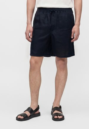 Homme portant un short noir à cordon et des sandales noires, debout devant un fond clair uni.
