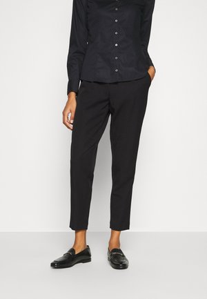 Person wearing black button-up shirt, black fitted ankle-length trousers, and black leather loafers, standing with one hand in pocket.