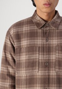 Camicia a quadri marrone con design a bottoni, due tasche frontali e un tessuto morbido e texture con colletto.
