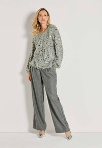 Eine Frau trägt einen silbernen Pailletten-Pullover mit Fuzzy-Effekt, graue, weit geschnittene, gestreifte Hosen, silberne Absätze und große Ohrringe, während sie vor einer weißen Wand steht.