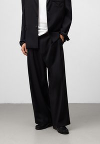 Svart skräddarsydd kavaj med en ficka och knäppning med en knapp, tillsammans med vida svarta byxor. Slitstukturerat tyg och välsittande passform. Svarta skor.