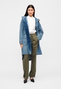 Μακρύ τζιν παλτό με κουκούλα, σε αποχρώσεις του πλενόμενου μπλε. Συνδυασμένο με λευκό πουκάμισο και ελαφρώς στενές παντελόνες σε ελιά πράσινη απόχρωση. Μαύρες μυτερές γόβες.