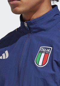 Marineblå vindjakke med høj krave, sølv lynlås og hvid branding. Har et broderet Italien FIGC-logo med grønne, hvide og røde detaljer.