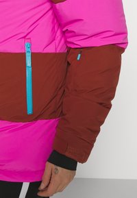 Doudoune avec des rayures horizontales roses et brunes, accentuée par une fermeture éclair bleue, en tissu texturé et dotée d'une sangle Velcro sur la manche.