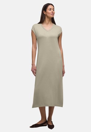 Kvinde iført en ærmeløs, beige midi-kjole med V-udskæring, kombineret med mørkebrune flade sko, stående mod en ensartet lys baggrund.