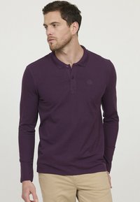 Polo à manches longues de couleur violet foncé, fabriqué en tissu doux. Il possède un col, une patte de boutonnage à deux boutons et un petit logo sur la poitrine.